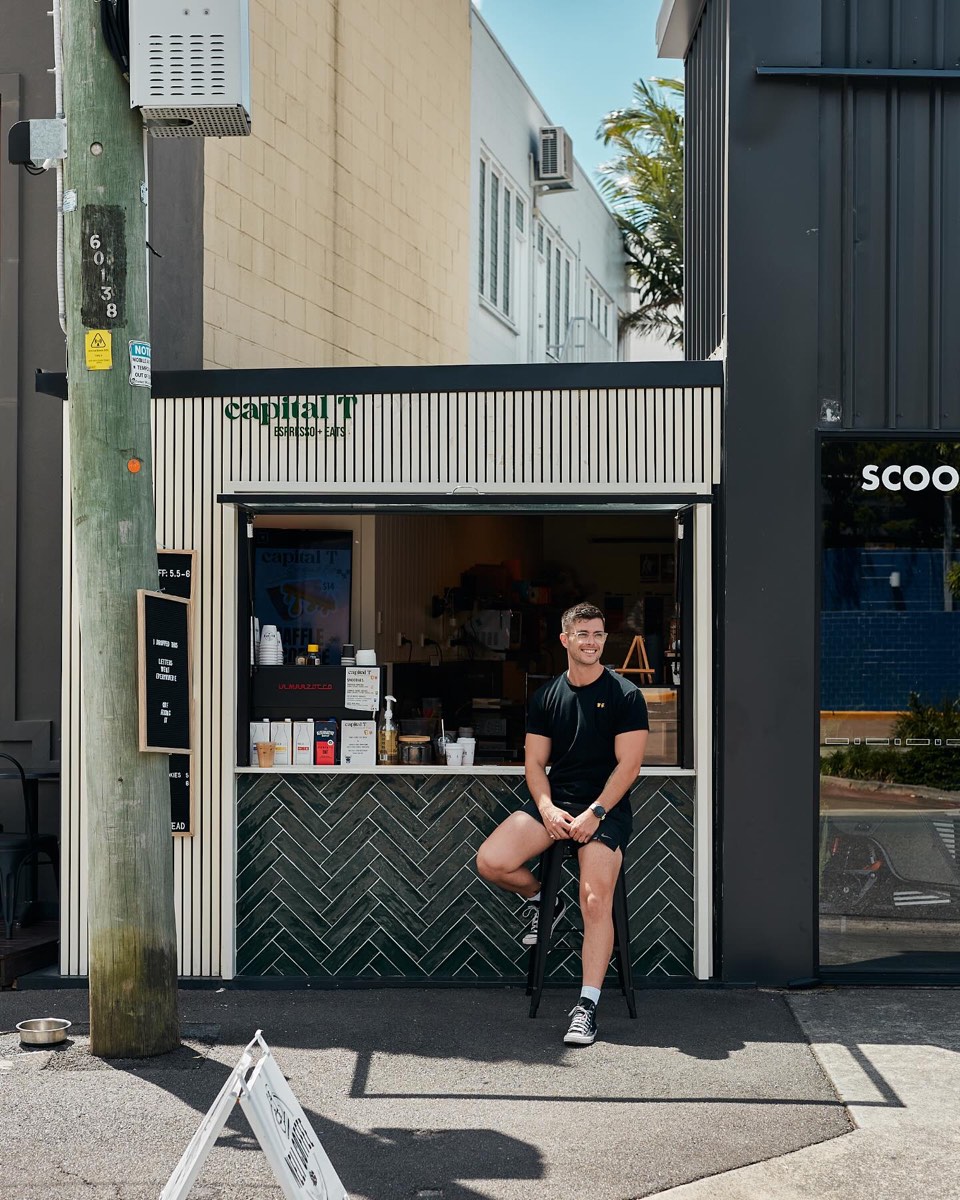 Tim Bates sitting on a stool outside the Capital T window, a cream-slatted shopfront with dark green herringbone tiles on Waterloo Street.